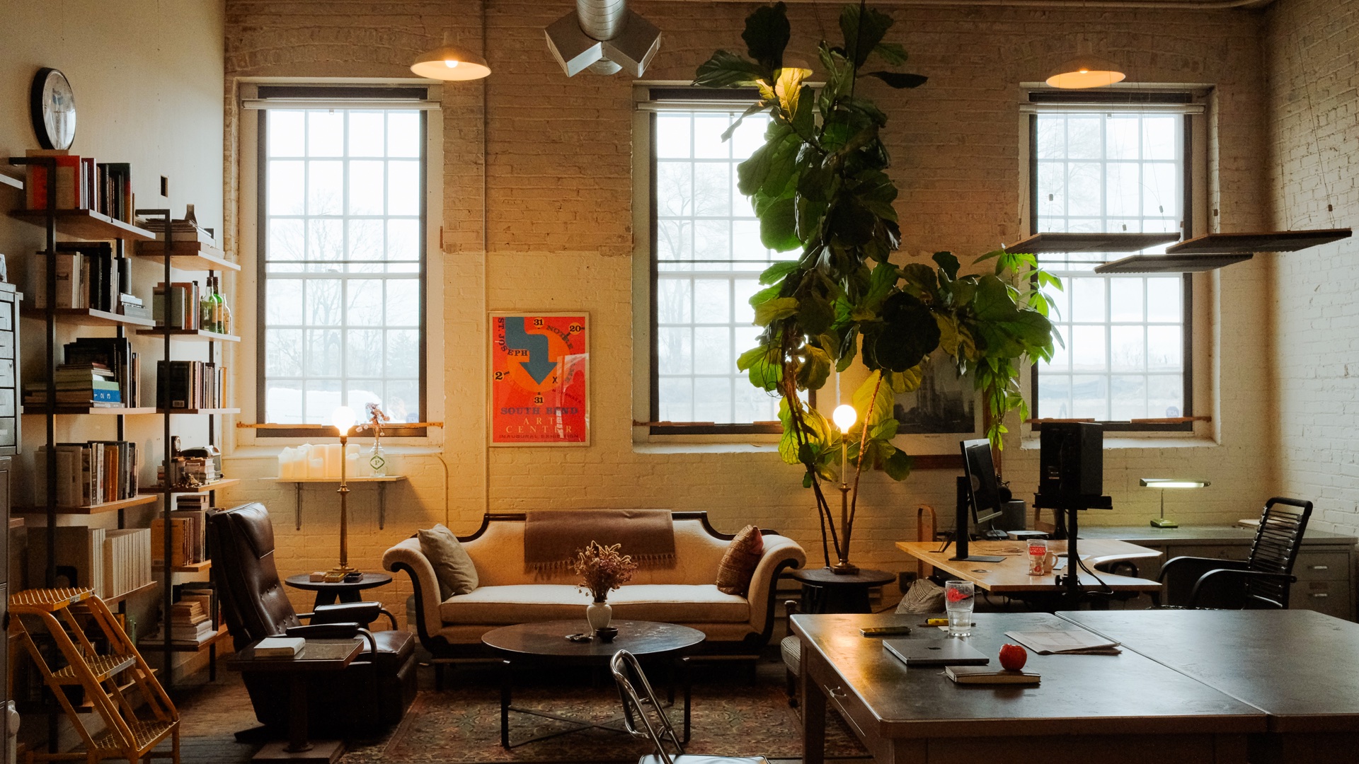 Interior studio at Vested Interest with tall factory windows, brick walls, bookshelves, desks, and lounge seating.